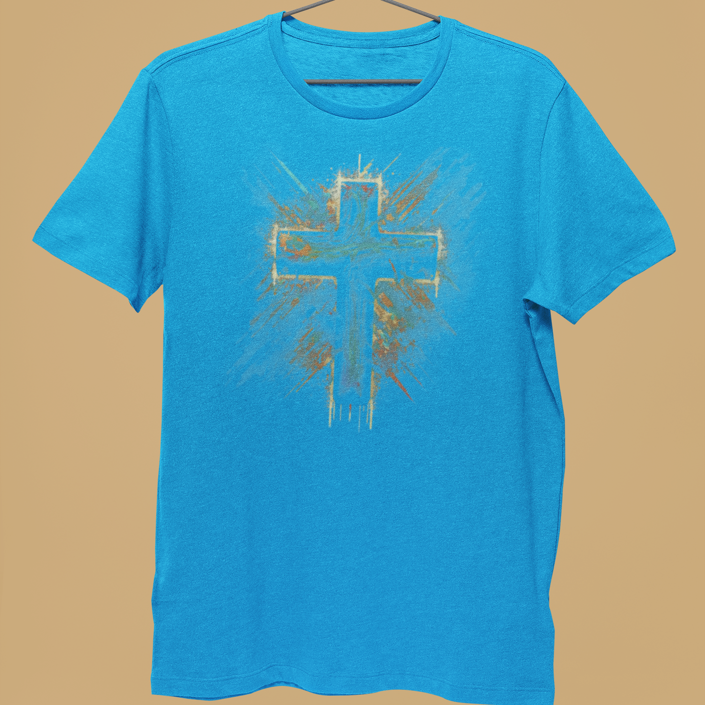 A bright blue short-sleeve t-shirt hanging on a metal hanger, held by a person's hand against a solid tan background. The shirt features a distressed, spray-painted style graphic of a cross in the center with gold and white paint details.