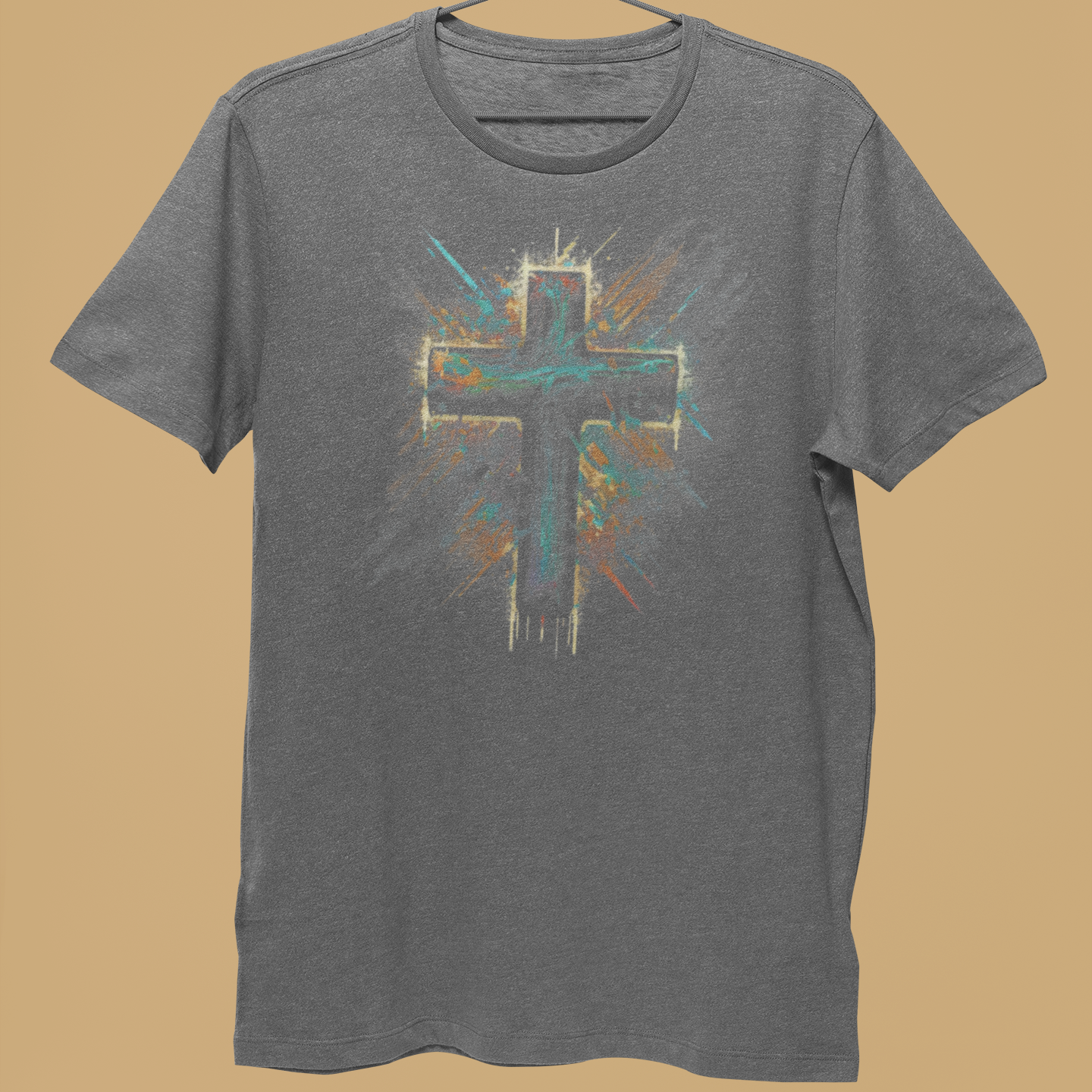 A heather gray short-sleeve t-shirt hanging on a metal hanger, held by a person's hand against a solid tan background. The shirt features a distressed graphic of a cross with teal, orange, and white paint splatter details.