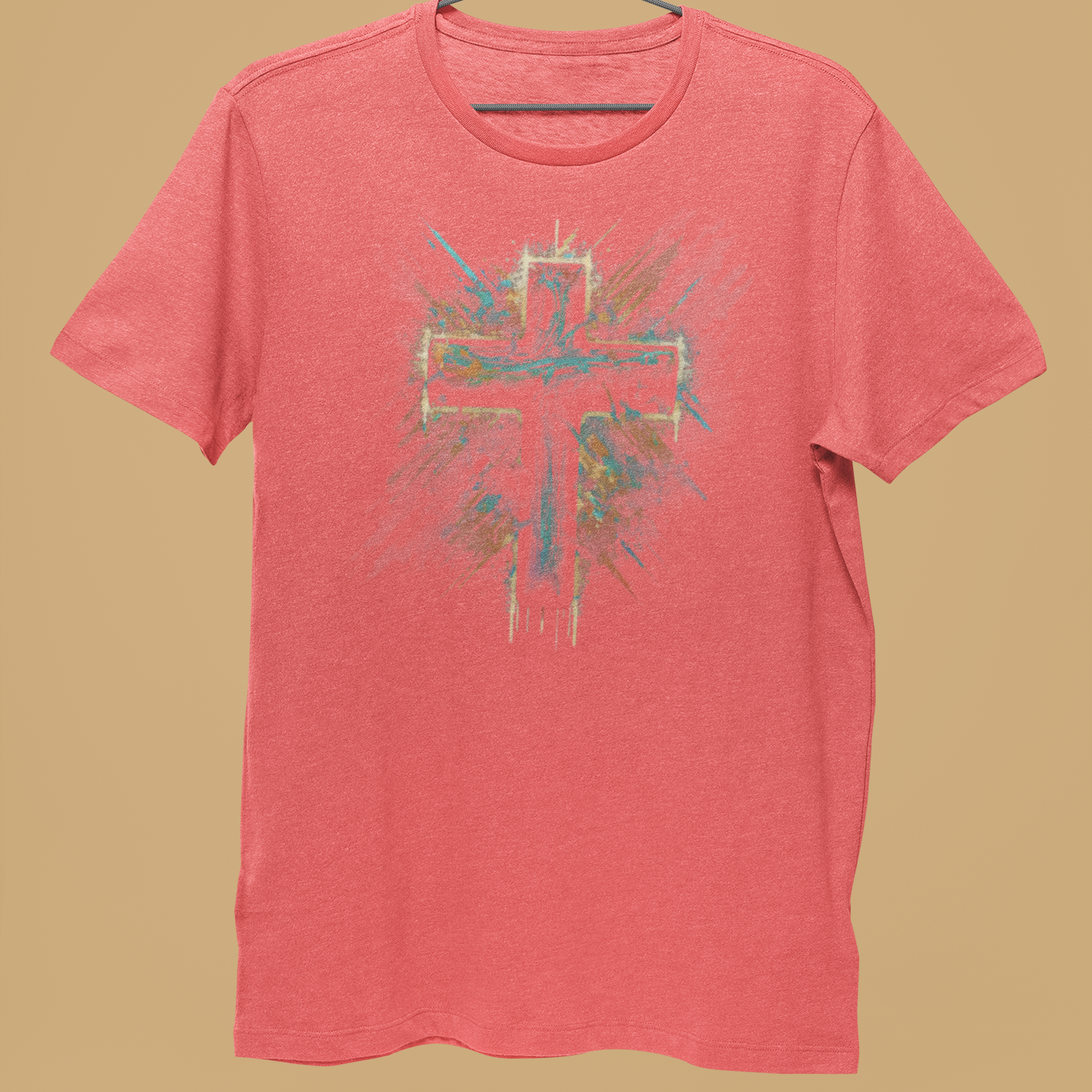 A coral pink heather t-shirt with a colorful cross graphic on the front, hanging on a black wire hanger held by a person's hand against a solid tan background.