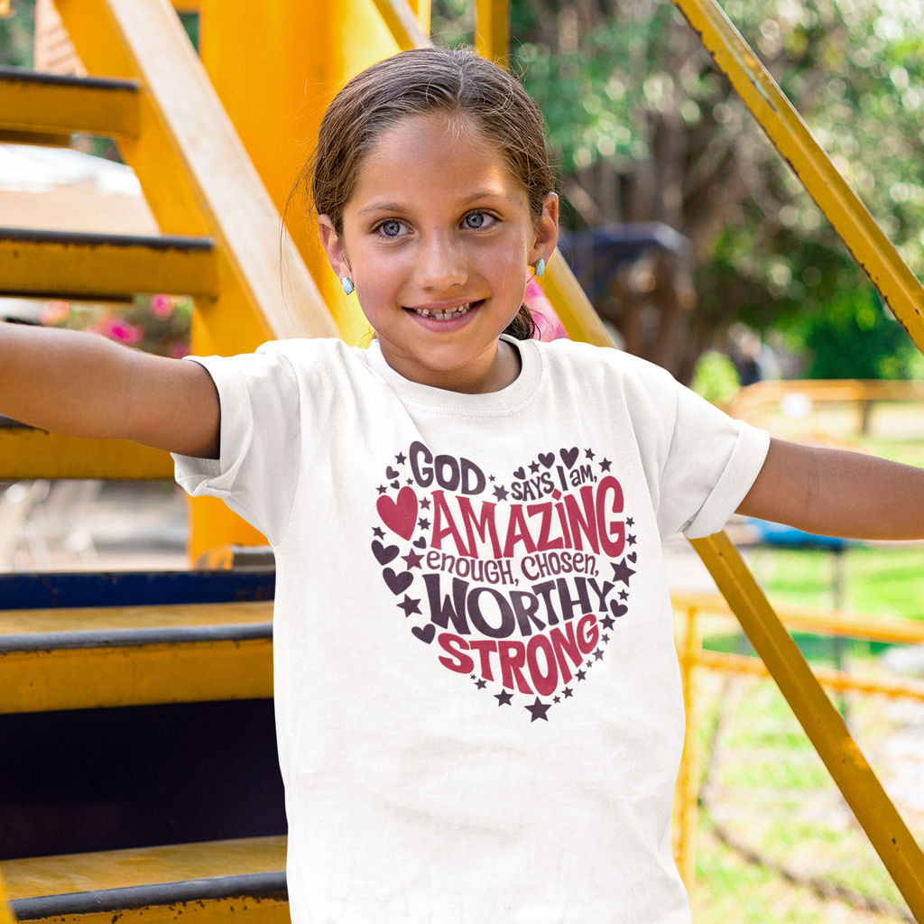 A young girl smiling while wearing a white graphic t-shirt with a red and black heart design that reads 'GOD IS MY AMAZING Caring WORTHY STRONG!'. She is standing on yellow playground stairs outdoors.
