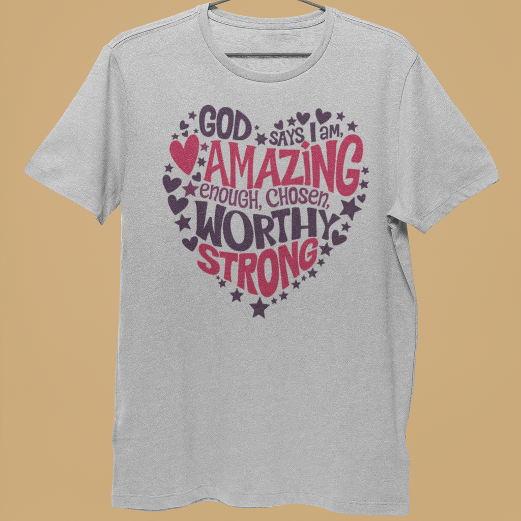 A light gray heather t-shirt hanging on a metal hanger held by a person's hand, featuring a heart-shaped graphic with the words 'GOD says I am AMAZING enough, CHOSEN WORTHY STRONG' in pink and dark purple lettering with stars and hearts, against a solid tan background.