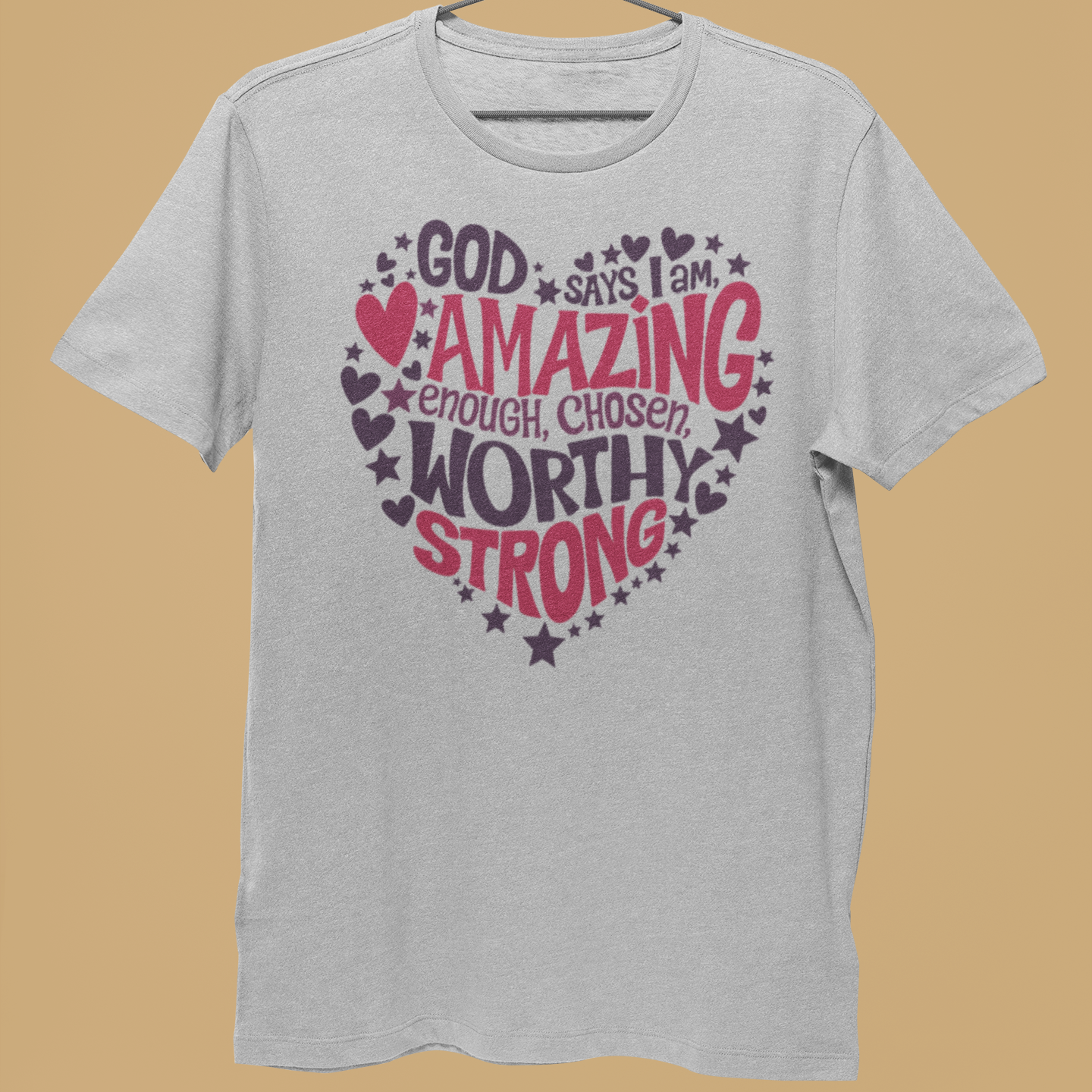 A light gray heather t-shirt hanging on a metal hanger held by a person's hand, featuring a heart-shaped graphic with the words 'GOD says I am AMAZING enough, CHOSEN WORTHY STRONG' in pink and dark purple lettering with stars and hearts, against a solid tan background.