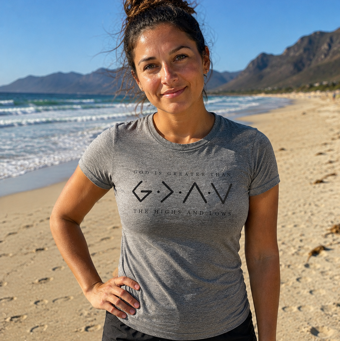 A woman standing on a sandy beach wearing a heather gray t-shirt with a black graphic print that reads 'G•A•V THE RISES AND THE LOWS'.