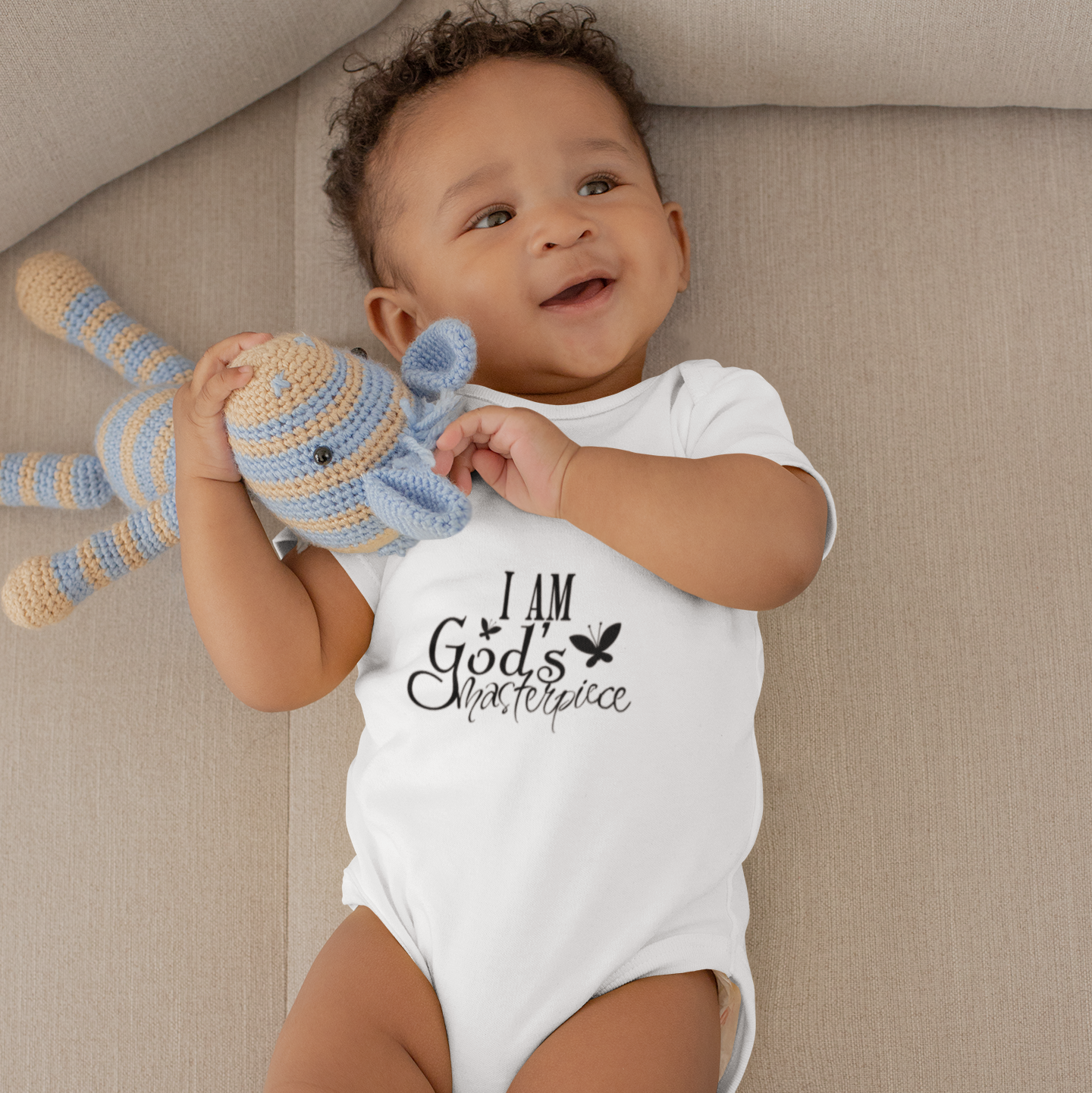 Baby on a beige couch wearing a white short sleeve baby grow printed with I Am God's Masterpiece.