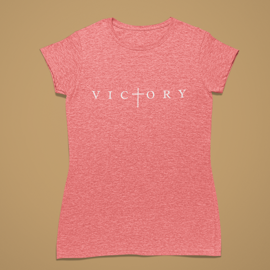 A heather pink short-sleeve t-shirt with the word 'VICTORY' printed in white across the chest, featuring a cross symbol where the 'I' is replaced. The shirt is displayed flat against a solid tan background.