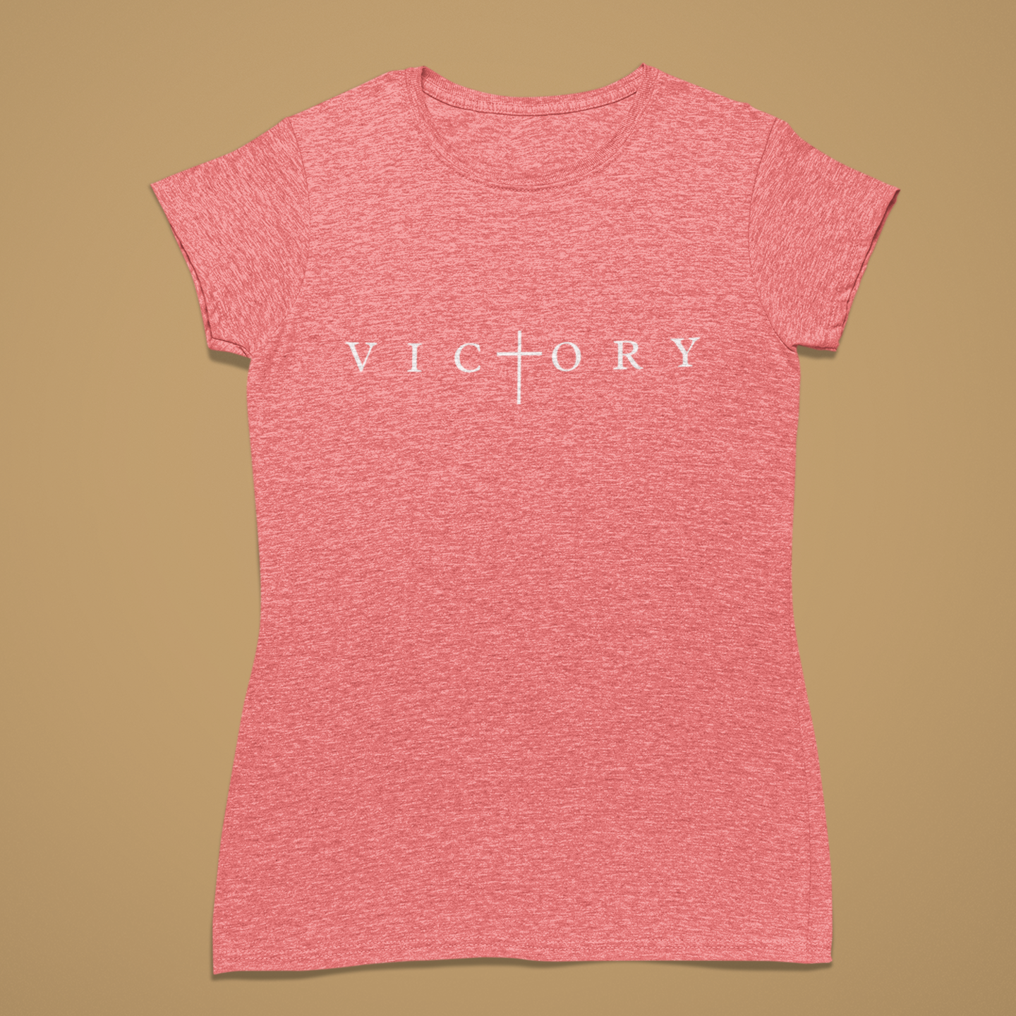 A heather pink short-sleeve t-shirt with the word 'VICTORY' printed in white across the chest, featuring a cross symbol where the 'I' is replaced. The shirt is displayed flat against a solid tan background.