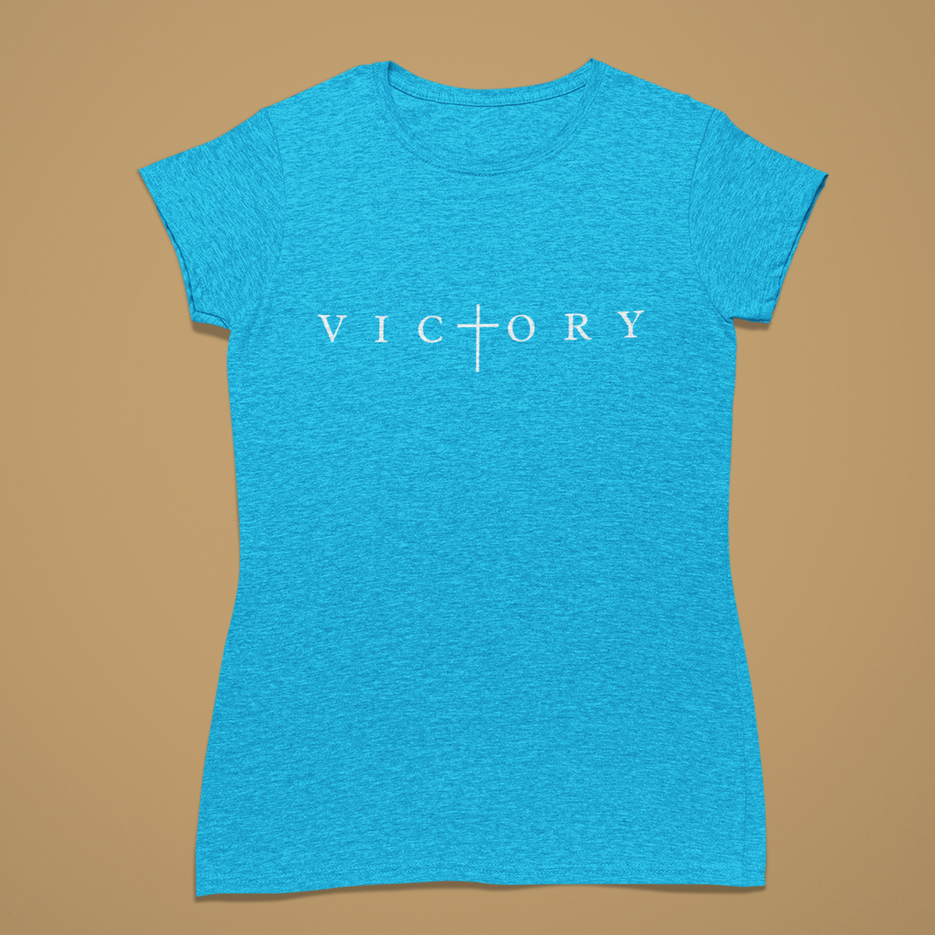 A women's short-sleeve crewneck t-shirt in heather blue, featuring the word 'VICTORY' printed in white across the chest with a cross symbol replacing the letter 'I'. The shirt is displayed flat against a solid tan background.