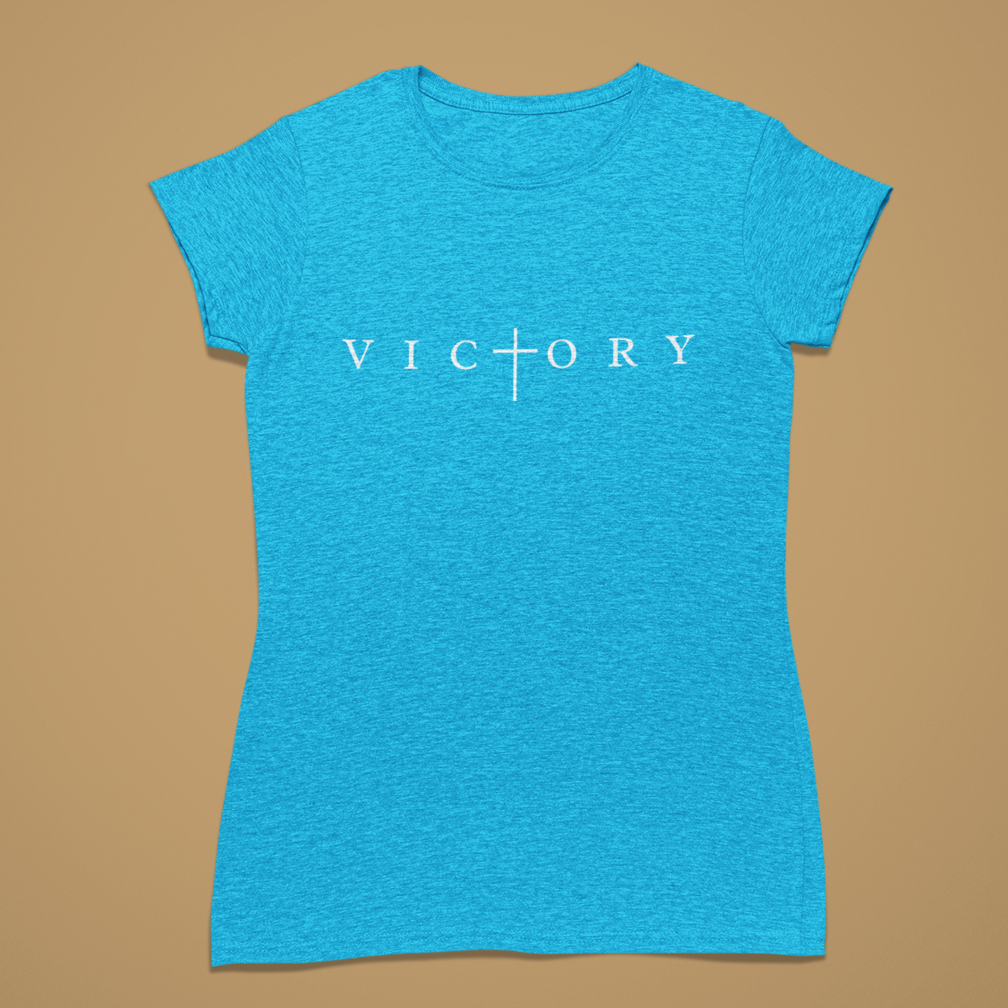 A women's short-sleeve crewneck t-shirt in heather blue, featuring the word 'VICTORY' printed in white across the chest with a cross symbol replacing the letter 'I'. The shirt is displayed flat against a solid tan background.