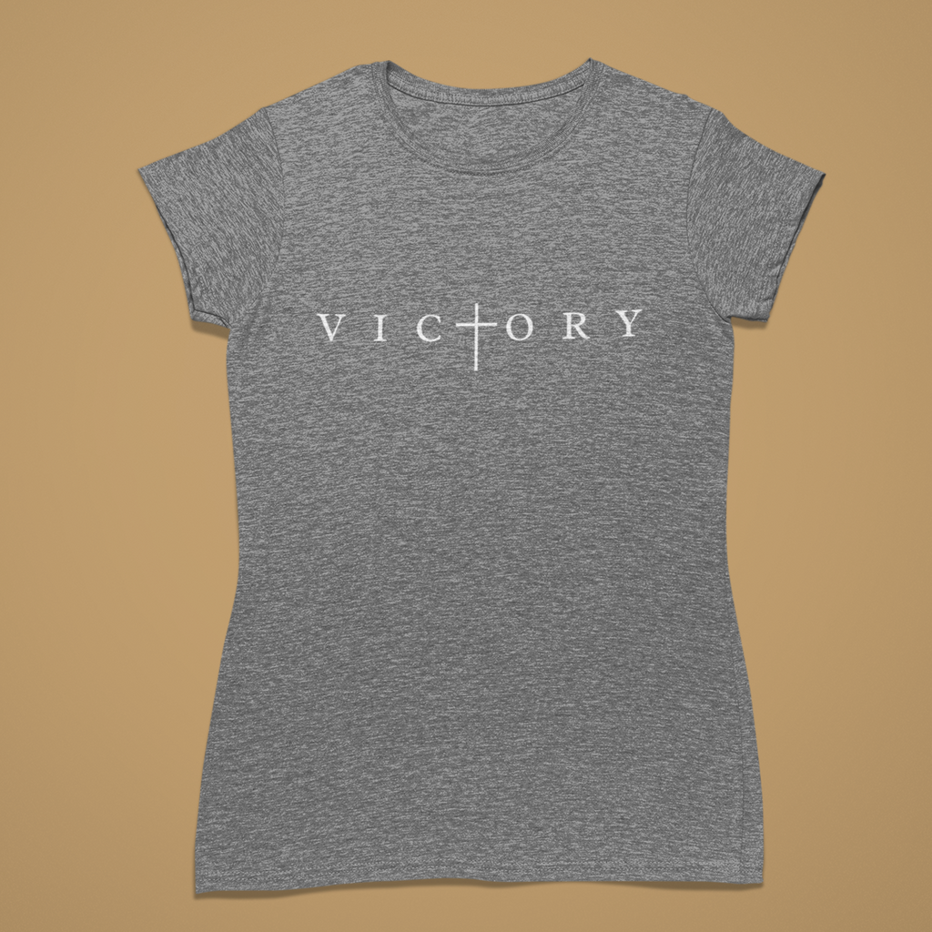 A women's short-sleeve crewneck t-shirt in heather grey, featuring the word 'VICTORY' printed in white across the chest with a cross symbol replacing the letter 'I'. The shirt is displayed flat against a solid tan background.