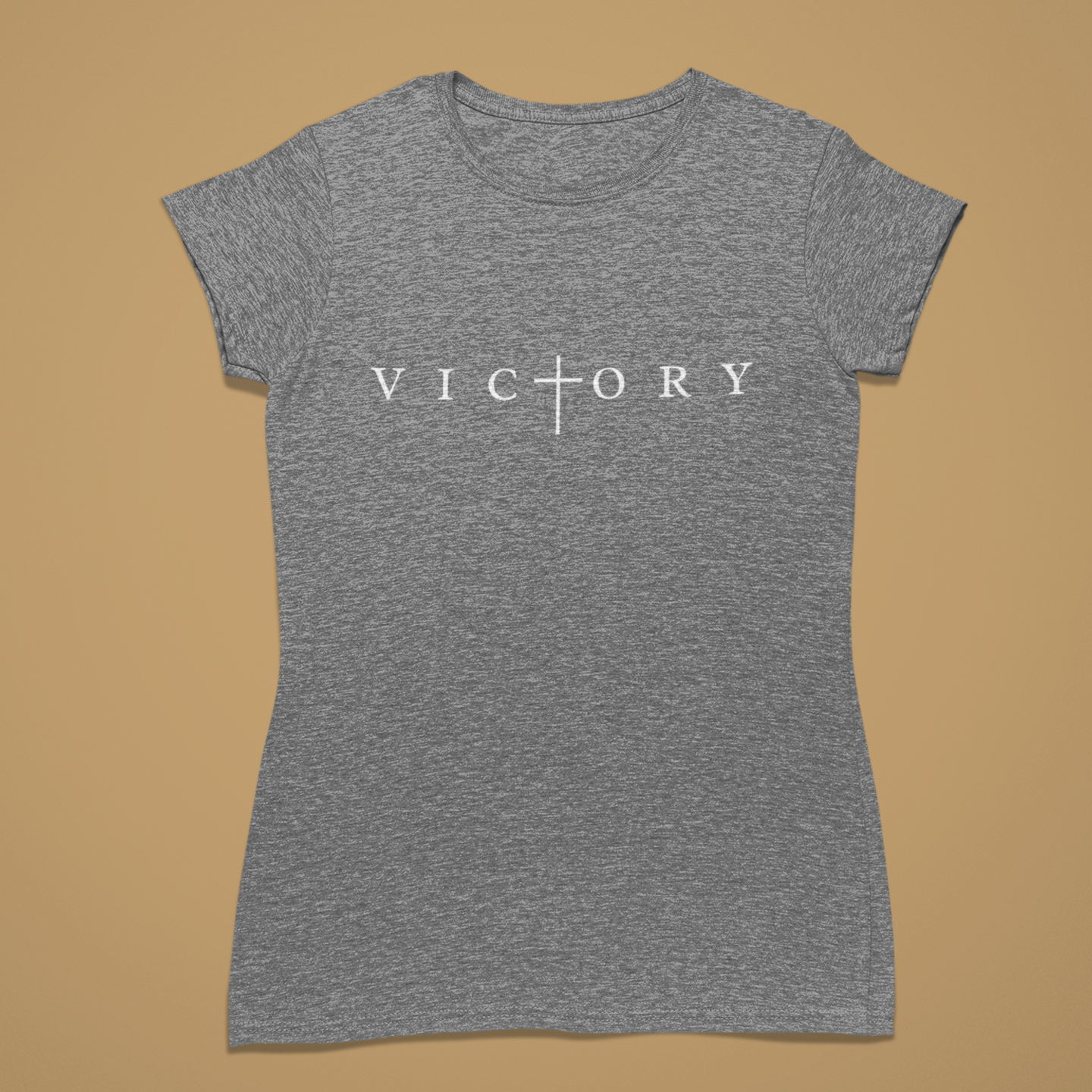 A women's short-sleeve crewneck t-shirt in heather grey, featuring the word 'VICTORY' printed in white across the chest with a cross symbol replacing the letter 'I'. The shirt is displayed flat against a solid tan background.