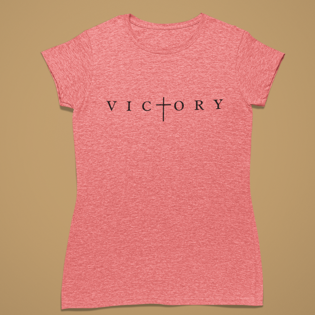 A heather pink short-sleeve t-shirt with the word 'VICTORY' printed in black across the chest, featuring a cross symbol where the 'I' is replaced. The shirt is displayed flat against a solid tan background.