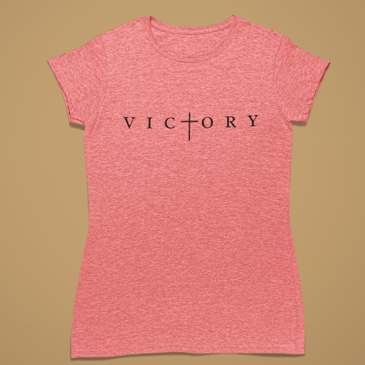 A heather pink short-sleeve t-shirt with the word 'VICTORY' printed in black across the chest, featuring a cross symbol where the 'I' is replaced. The shirt is displayed flat against a solid tan background.