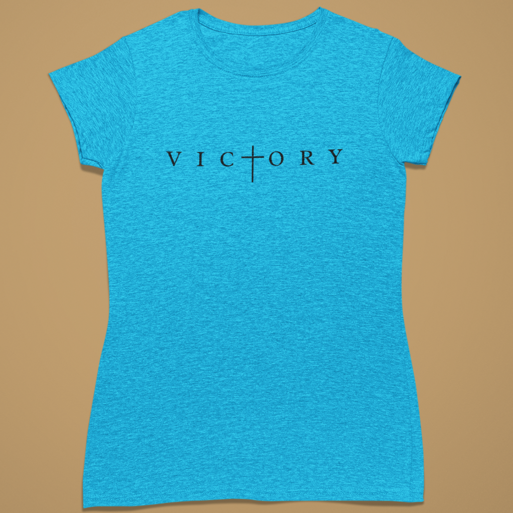 A women's short-sleeve crewneck t-shirt in heathered bright blue, featuring the word 'VICTORY' printed across the chest in black serif letters with a cross symbol replacing the 'I'. The shirt is displayed flat against a solid tan background.