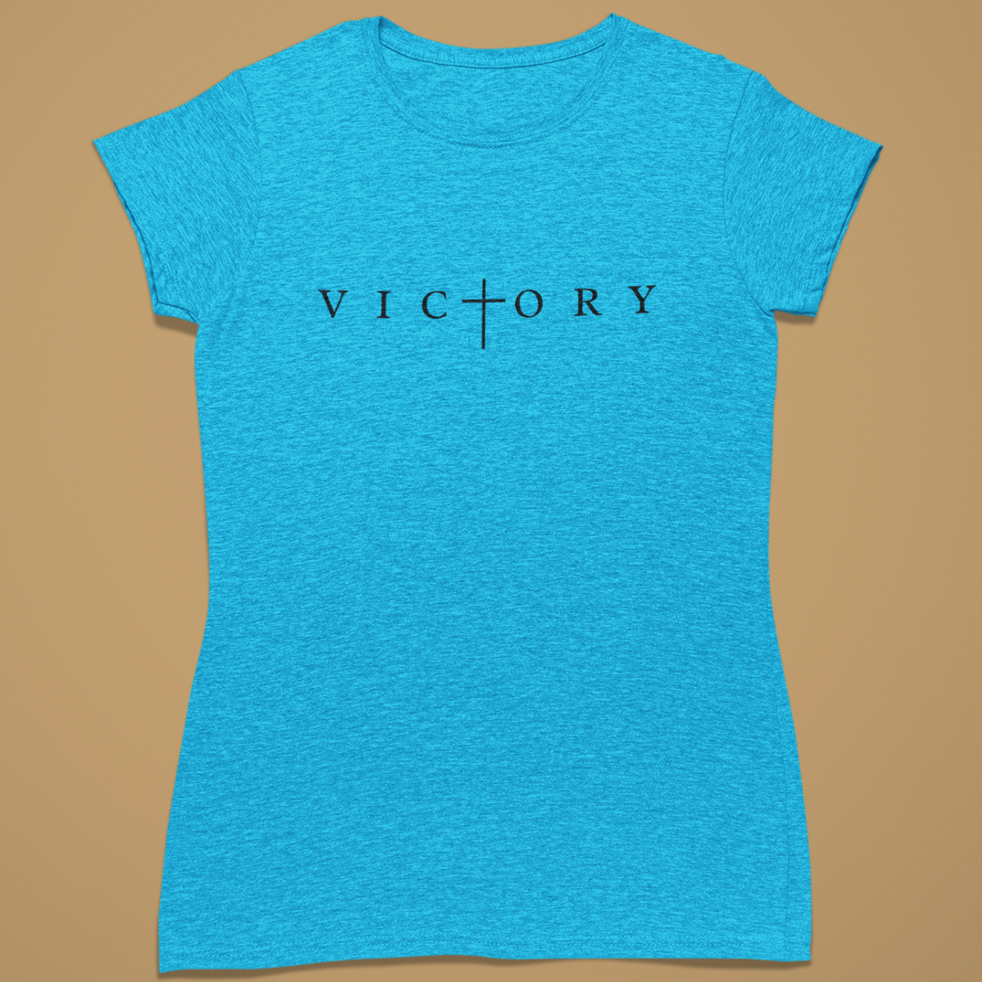 A women's short-sleeve crewneck t-shirt in heathered bright blue, featuring the word 'VICTORY' printed across the chest in black serif letters with a cross symbol replacing the 'I'. The shirt is displayed flat against a solid tan background.