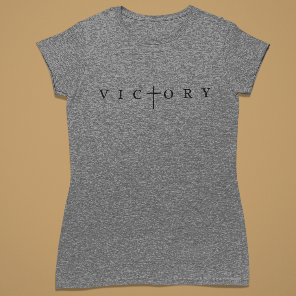 A heather gray short-sleeve t-shirt with the word 'VICTORY' printed across the chest in black, where the letter 'T' is replaced by a cross symbol. The shirt is displayed flat against a solid tan background.