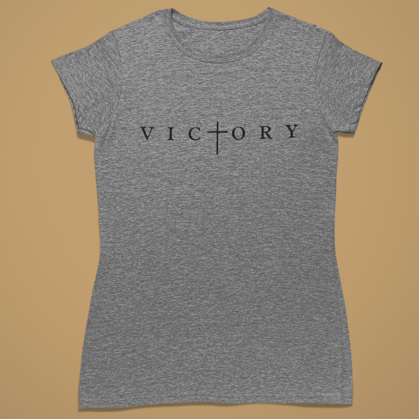 A heather gray short-sleeve t-shirt with the word 'VICTORY' printed across the chest in black, where the letter 'T' is replaced by a cross symbol. The shirt is displayed flat against a solid tan background.
