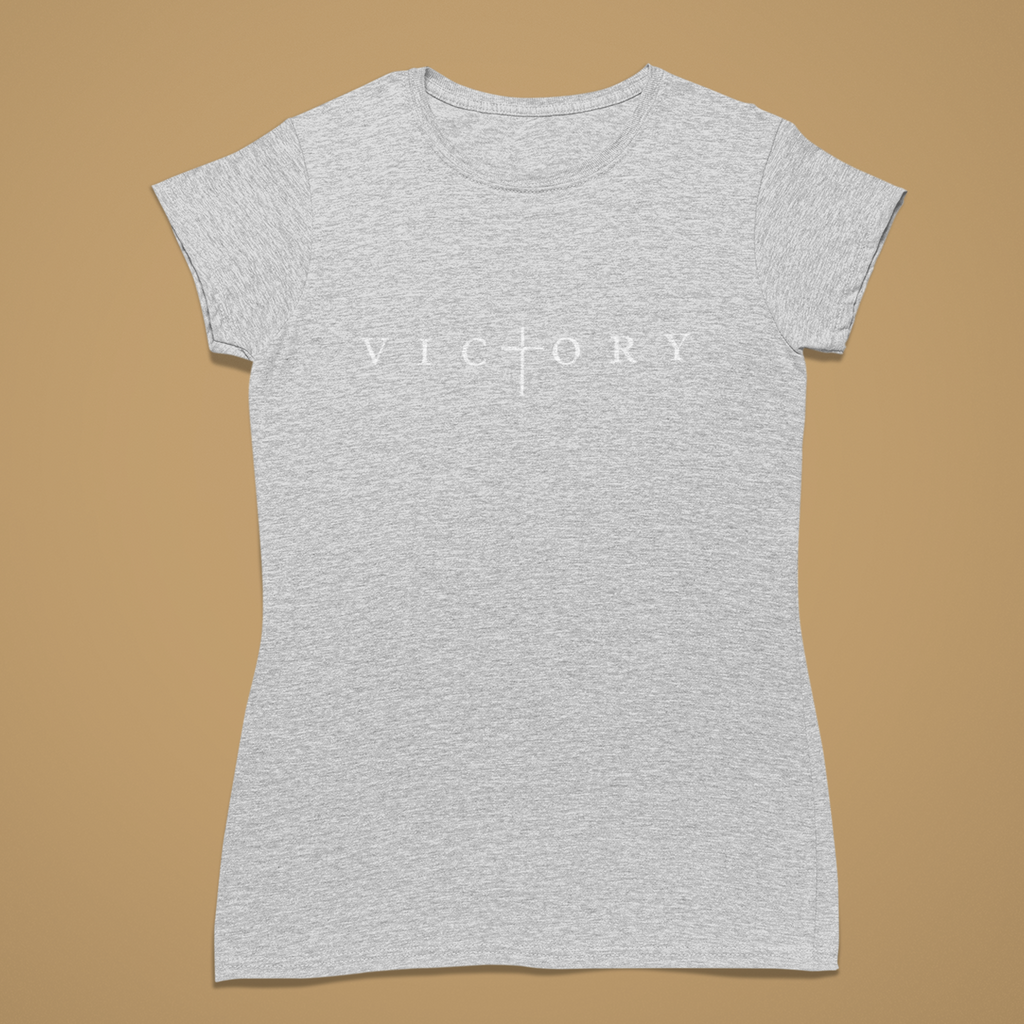 A women's short-sleeve crewneck t-shirt in heather light grey, featuring the word 'VICTORY' printed in white across the chest with a cross symbol replacing the letter 'I'. The shirt is displayed flat against a solid tan background.