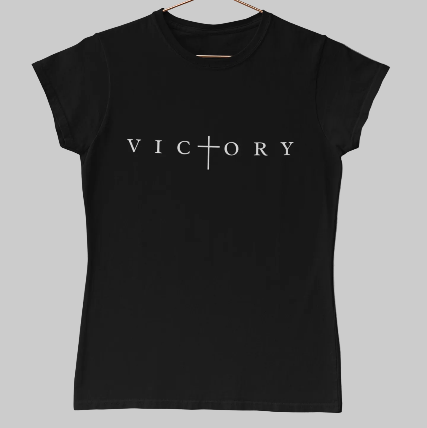 A women's short-sleeve crewneck t-shirt in solid black, featuring the word 'VICTORY' printed in white across the chest with a cross symbol replacing the letter 'I'. The shirt is displayed flat against a solid concrete background.