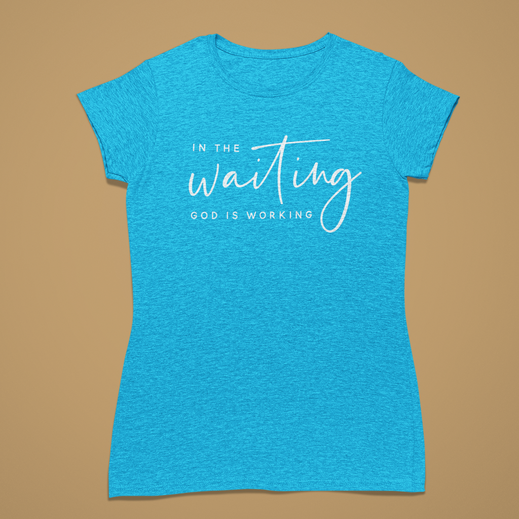 A heather blue short-sleeve t-shirt with a white graphic text print reading 'IN THE waiting GOD IS WORKING' on the front, displayed against a solid tan background.
