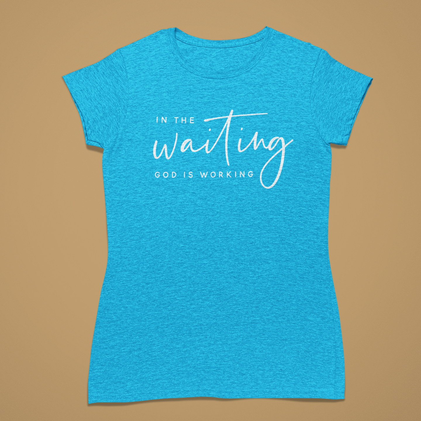 A heather blue short-sleeve t-shirt with a white graphic text print reading 'IN THE waiting GOD IS WORKING' on the front, displayed against a solid tan background.
