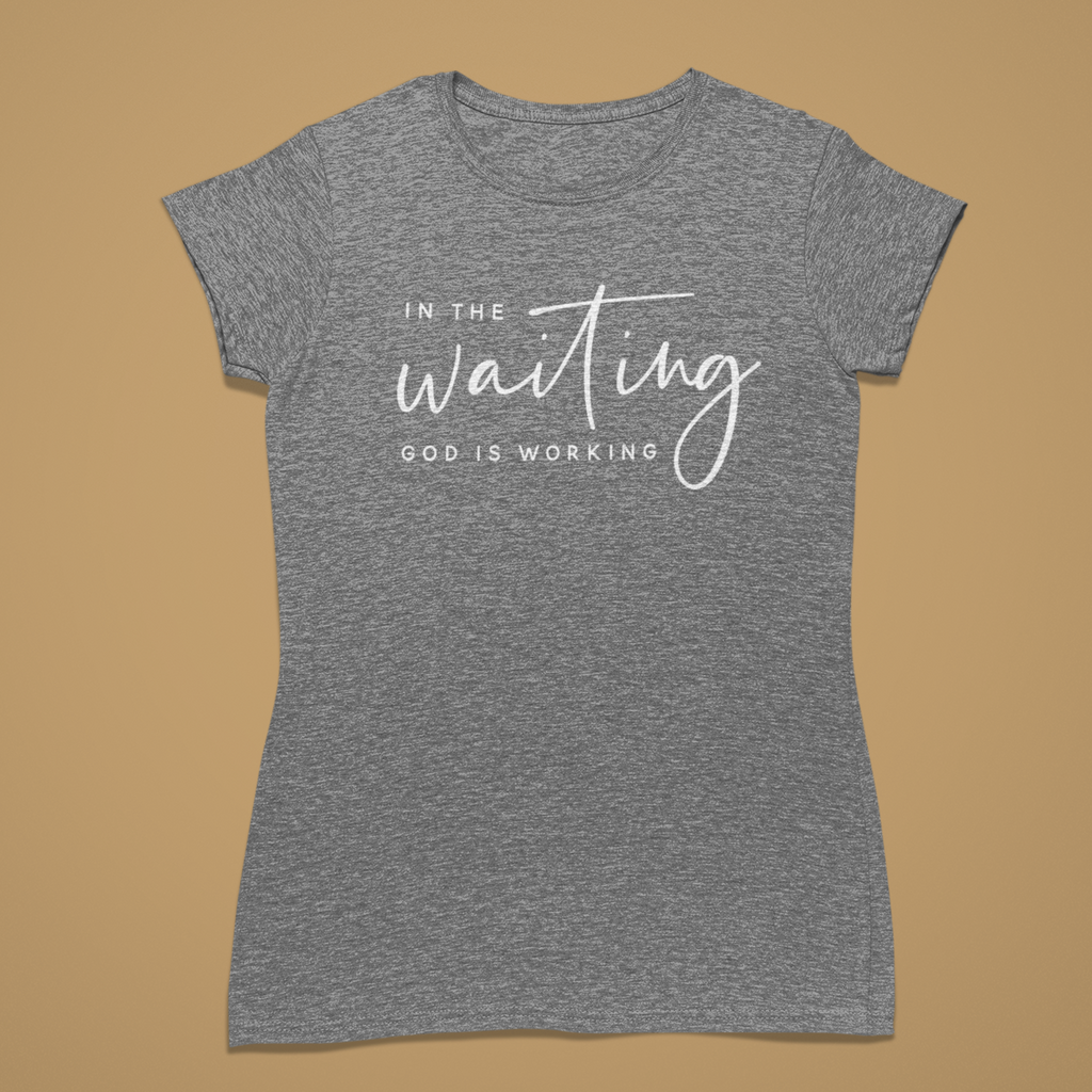 A heather gray short-sleeve t-shirt with a white graphic text print reading 'IN THE waiting GOD IS WORKING' on the front, displayed flat against a solid tan background.