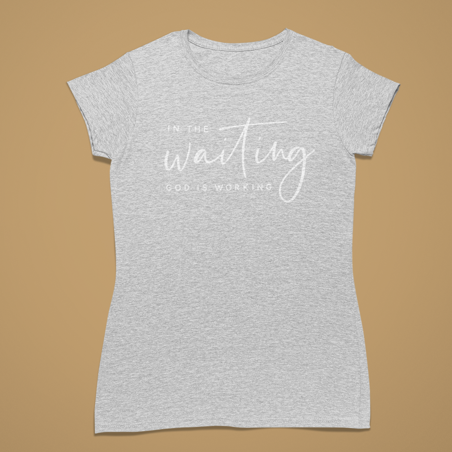 A light heather gray short-sleeve t-shirt with white printed text reading 'IN THE waiting GOD IS WORKING' on the front, displayed flat against a solid tan background.
