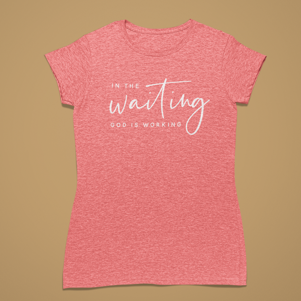 A heather pink short-sleeve t-shirt with white printed text reading 'IN THE waiting GOD IS WORKING' on the front, displayed flat against a solid tan background.
