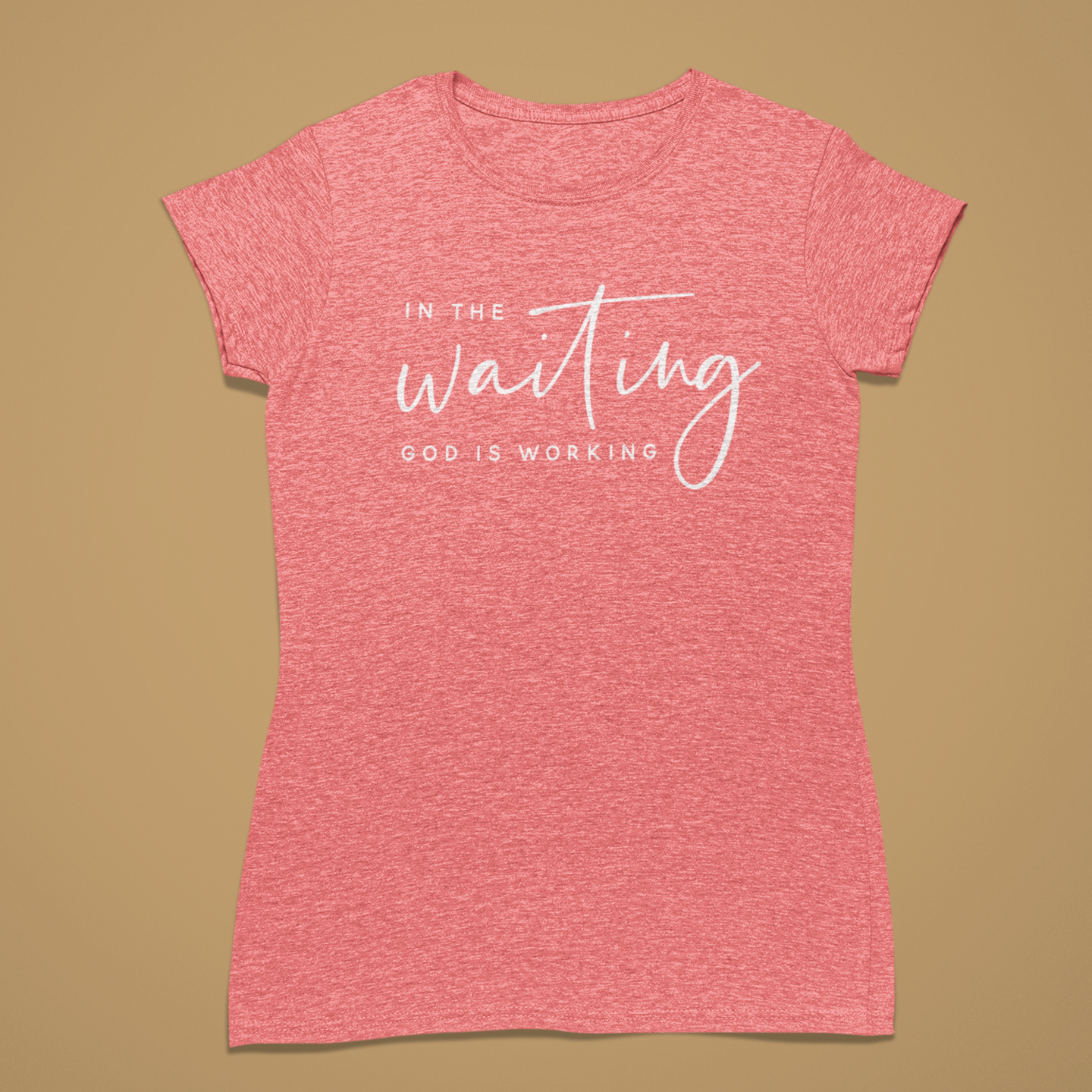 A heather pink short-sleeve t-shirt with white printed text reading 'IN THE waiting GOD IS WORKING' on the front, displayed flat against a solid tan background.